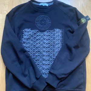 Svart Stone Island sweatshirt L - Svart sweatshirt från Stone Island i slim fit med broderad logga på bröstet och unikt grafiskt mönster framtill. Klassisk rund hals, ribbade muddar och ikonisk patch på ärmen. Perfekt för dig som gillar streetwear och snygga detaljer.