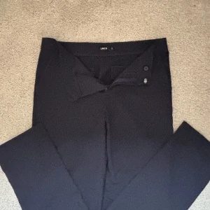 Svarta kostymbyxor från LMTD S/M - Stilrena svarta kostymbyxor från LMTD i klassisk rak modell. Byxorna har diskreta bakfickor, bälteshällor och stängs med knapp och dragkedja. Perfekta för en clean och dressad look.