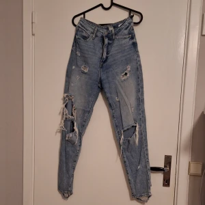 Ljusblå ripped jeans med hög midja - Säljer ett par ljusblå jeans med hög midja och rejält slitna detaljer på benen. Jeansen har smal passform och är tillverkade i klassiskt denimtyg. Perfekta för dig som gillar en edgy och avslappnad stil.