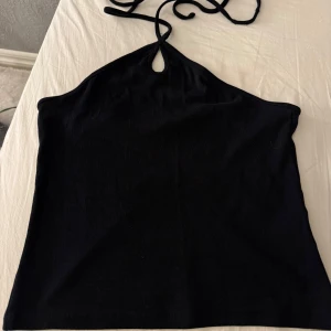 Svart halterneck linne med knyt - Säljer en svart halterneck linne med smala knytband i nacken och en liten droppformad öppning framtill. Toppen är ärmlös och har en enkel, stilren design som passar perfekt till jeans eller kjol. Mjuk och stretchig bomullskänsla. Det enda problemet är att etiketten är väldigt lös på ena sidan vilket orsakat två jätte små hål i baksidan som inte alls är synliga. 
