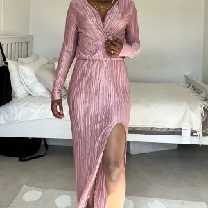 Ljusrosa långklänning med slits - Säljer en snygg ljusrosa långklänning med plisserat tyg och v-ringad omlottfront. Klänningen har långa ärmar och en hög slits framtill som ger en cool vibe. Materialet är glansigt och känns mjukt mot huden, perfekt för dig som vill sticka ut.