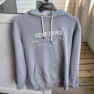 Grå hoodie med broderi från H&M - Säljer en chill grå hoodie från H&M med vit broderad text 'SELF LOVE' på bröstet. Hoodien har snörning i huvan, stor magficka och långa ärmar. Perfekt för dig som gillar comfy vibes och snygga detaljer. Passar dig som vill ha en enkel men ändå stilren look.