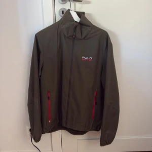 Olivgrön vindjacka från Polo - Snygg olivgrön vindjacka från Polo med röda dragkedjor och logga på bröstet. Jackan har hög krage, långa ärmar och två fickor framtill. Perfekt för blåsiga dagar och enkel att matcha med streetwear eller casual outfits.