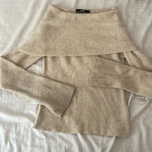 Beige offshoulder stickad tröja Bik Bok - Supermysig beige stickad tröja från Bik Bok i ullblandning. Offshoulder-modell med bred krage och långa ärmar. Ribbad struktur som ger en snygg och chill vibe. Perfekt att styla med jeans eller kjol för en trendig look.