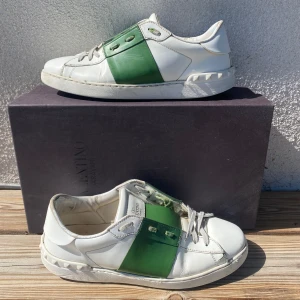 Valentino open - Snygga vita sneakers från Valentino Garavani med en bred grön läderdetalj över sidan. Skorna har rund tå, platt sula och klassisk snörning. Kommer med original dustbag och låda. Perfekt för dig som gillar exklusiva och stilrena sneakers. Liten defekt på högerskott där man ser en spricka men inget som förstör skorna eller annat. Skick 8/10