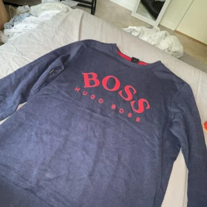 Blå tröja från Hugo Boss med röd logga - Snygg marinblå tröja från Hugo Boss i bomull med stor röd BOSS-logga framtill. Klassisk rund halsringning med röd detalj på insidan av kragen. Långärmad och passar perfekt till jeans eller joggers.