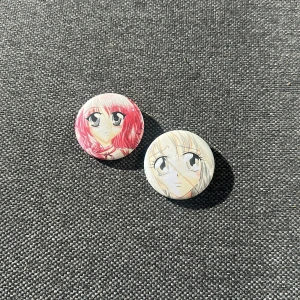 Anime-inspirerade pins med motiv - Två runda pins med handritade anime-motiv, en med rosa hår och en med blont hår. Kommer i en genomskinlig vit organzapåse med snörning. Perfekt att fästa på väskan eller jackan för att visa din stil. Metallbaksida med säkerhetsnål för enkel fastsättning.