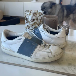 Valentino Garavani sneakers - Snygga sneakers från Valentino Garavani i vitt skinn med bred blå läderdetalj över snörningen. Klassisk rund tå, platt sula och diskret logga på plösen. Perfekta för dig som gillar exklusiva och stilrena sneakers med en unik touch.