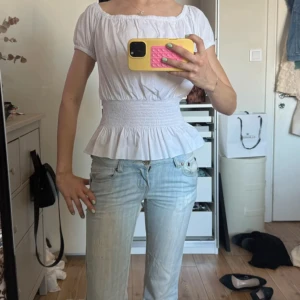 Vit blus med smock och snörning - Supersöt vit blus med smockad midja och volang nedtill. Toppen har korta ärmar, offshoulder-modell och snörning i ryggen som ger en snygg passform. Perfekt att styla med jeans för en fräsch look.