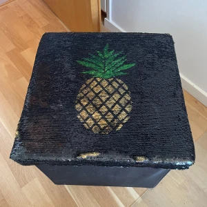 Cool sittpall/förvaringslåda - Bilderna visar en svart förvaringslåda med lock dekorerat med paljetter och en ananasdesign. Locket kan ändra utseende när man drar med handen över paljetterna. Lådan är rymlig och passar perfekt för att förvara saker i rummet. Ingen bok syns på bilderna.