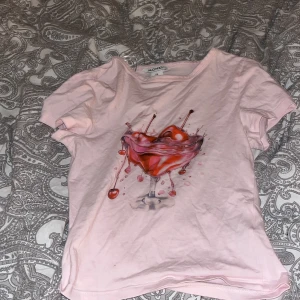 Rosa t-shirt med drinkmotiv från Monki - Snygg ljusrosa t-shirt från Monki med ett coolt tryck av en drink med körsbär på bröstet. T-shirten har rund hals och korta ärmar, perfekt för dig som gillar unika prints. 