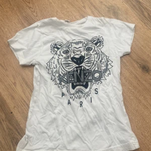 Vit Kenzo t-shirt med tigertryck - Snygg vit t-shirt från Kenzo med stort tigertryck och logga på bröstet. Rund hals och korta ärmar. Materialet är mjuk bomull som känns skönt mot huden. Perfekt för dig som gillar statement-plagg och coola prints.