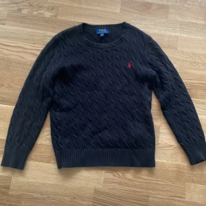 Svart stickad tröja Polo Ralph Lauren - Snygg svart stickad tröja från Polo Ralph Lauren med klassiskt kabelstickat mönster och röd logga, Stilren, mycket bra skick. Ser kanske mörkblå ut på bilderna men jag lovar att den är svart