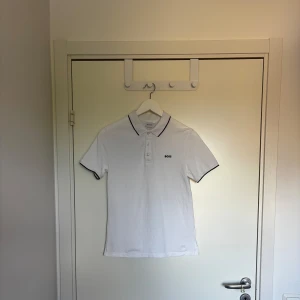 Vit BOSS piké med svarta detaljer - Vit pikétröja från BOSS med svart broderad logga på bröstet och svarta tunna kantdetaljer på krage och ärmslut. Klassisk krage och knappslå framtill. Tillverkad i mjuk bomull som känns skön mot huden. Perfekt för en clean och stilren look. Storleken är S i junior men är väldigt liten i storleken. Va inte rädda att höra av dig vid minsta lilla fundering😄