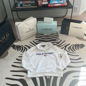 Vit half zip Polo Sport-tröja - Vit långärmad tröja från Polo Sport Ralph Lauren med blå och röd logga framtill och amerikansk flagga. Tröjan har en klassisk half zip-krage och är tillverkad i mjuk bomull, perfekt för en sportig och clean look.