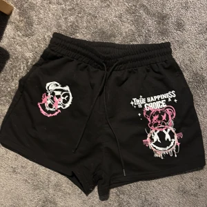 Svarta shorts med graffiti-tryck - Svarta shorts med coola graffiti-inspirerade tryck i vitt och rosa, samt texten 'True Happiness is a Choice'. De har snörning i midjan och en loose passform, perfekta för chill dagar. Materialet känns som mjuk bomull.