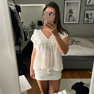 Neo noir blus - Supersöt vit blus från Neo Noir med v-ringning, volangdetaljer och lätt transparent tyg. Blusen har korta ärmar och ett luftigt, flowy fall som ger en romantisk vibe. Perfekt att styla med kjol eller jeans för en fräsch look.
