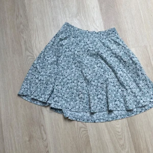 Blommig grå kortkjol XS/S - Supersöt grön kortkjol med små vita och svarta blommor. Kjolen har en mjuk och stretchig resår i midjan och är gjord i ett lätt bomullsmaterial. Perfekt för dig som gillar blommigt och vill ha en skön kjol med flowig passform.