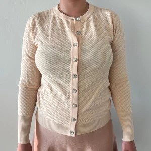 Beige/rosa kofta med pärlknappar - Snygg beige kofta med långärmad passform och glittriga pärlknappar framtill. Koftan har ett diskret strukturerat mönster och ribbade muddar vid ärmslut och nederkant. Perfekt för dig som gillar stilrena och mjuka plagg.