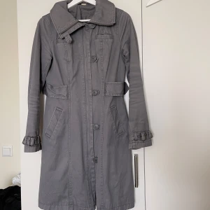 Grå trenchcoat med bältdetaljer - Snygg grå trenchcoat med stora knappar och bältdetaljer vid ärmsluten. Jackan har klassisk krage, två fickor framtill och markerad midja. Perfekt för dig som gillar stilrena och tidlösa plagg. Långärmad och i mjukt bomullsmaterial.