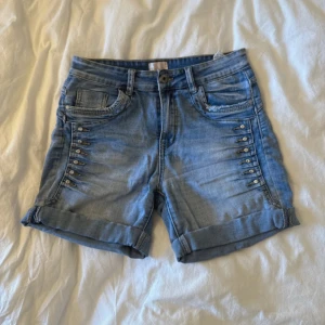 Blå jeansshorts med nitar och uppvik - Snygga blå jeansshorts med uppvikta ben och coola nitar längs sidorna. Klassisk femficksmodell med knapp och dragkedja framtill. Baksidan har två fickor, varav en med en liten stjärndetalj. Perfekta för sommarens alla häng.