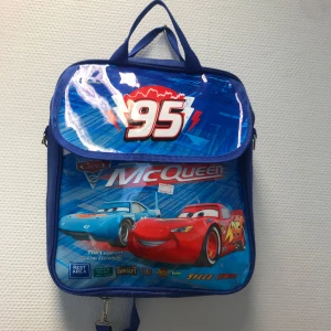 Blå Cars ryggsäck - Blå fyrkantig ryggsäck med motiv Cars . Stor 95-logga på locket och färgglada tryck med bilar på framsidan. Väskan har justerbara axelremmar och bärhandtag. Tillverkad i slitstark polyester, perfekt för skolan eller utflykter.