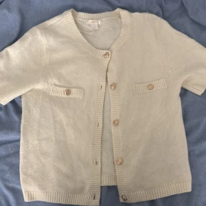 Beige kofta från Gina Tricot med knappar - Kortärmad beige kofta från Gina Tricot med snygga guldfärgade knappar framtill och på de två dekorativa fickorna. Koftan är stickad med ribbade kanter vid ärmslut, halsringning och nederkant. Perfekt för lager-på-lager och enkel att styla.