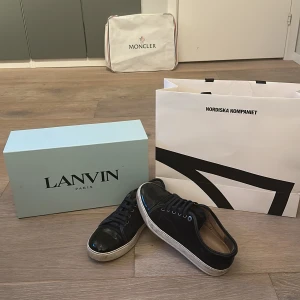 Svarta sneakers från Lanvin - Säljer ett par svarta sneakers från Lanvin med blanka detaljer på tån och snörning framtill. Skorna har vit sula och är tillverkade i en kombination av skinn och syntet. Klassisk och stilren design som passar till mycket.