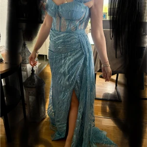 Ljusblå glittrig långklänning med slits - Säljer en ljusblå långklänning med glittrigt mönster och markerad midja. Klänningen har smala axelband, transparenta partier upptill och en hög slits framtill. Perfekt för bal eller fest! 💙