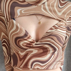 Brun mönstrad croppad topp med cut-out - Säljer en croppad topp med brunt, beige och vitt vågmönster. Toppen har lång ärm, hög hals och en snygg cut-out detalj framtill. Materialet är stretchigt och känns mjukt mot huden. Den är rätt så oanvänd, kanske använt den 2 gånger, lapparna är bortklippta då jag klipper bort de då jag tycker det klias så jag vet inte riktigt vilken storlek det är men jag som har s får på den, men skulle gissa apå att dne är xs