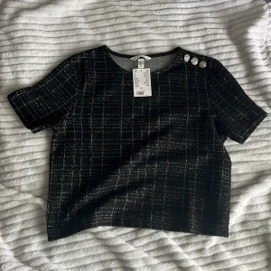 Svart topp med silvriga knappar från H&M  - Säljer en svart topp från H&M med rutigt mönster i vit tråd. Toppen har korta ärmar och tre dekorativa silvriga knappar på ena axeln. Materialet är strukturerat och ger en exklusiv känsla. Inte använd. ✨