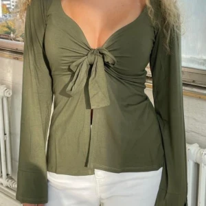 Olivgrön långärmad topp med knyt - Trendig olivgrön topp med lång ärm och djup v-ringning. Snygg knytdetalj framtill som ger en feminin touch. Perfekt att styla med jeans eller kjol för en cool look.