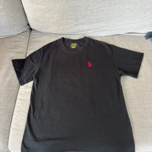 Svart t-shirt från Polo Ralph Lauren - Säljer en svart t-shirt från Polo Ralph Lauren med röd broderad logga på bröstet. Modellen är custom slim fit och har en klassisk rund halsringning. Perfekt basplagg med stilren design. Skick 8/10 Nypris 1099 mitt pris 300! PASSAR 166-178