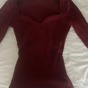 Vinröd ribbad topp  - Säljer en vinröd ribbad topp  med lång ärm och snygg v-ringad halsringning. Toppen har en tight passform och är perfekt att styla till olika outfits.