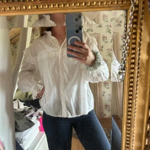 Vit blus med spets och volang - Säljer en romantisk vit blus med hög krage, spetsdetaljer och volanger på axlar och ärmslut. Blusen har knappar framtill och är långärmad. Perfekt för dig som gillar vintageinspirerad stil!