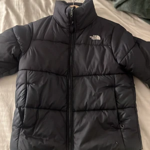 The North Face jacka - Säljer en svart dunjacka från The North Face med hög krage och dragkedja framtill. Jackan har lång ärm och klassisk pufferdesign med diskret logga på bröstet och ryggen. Perfekt för kyliga dagar.