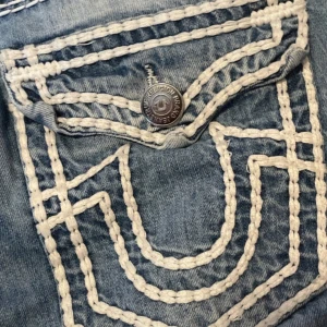 True Religion jeans med vit kontrastsöm - Säljer ett par blå jeans från True Religion med tydliga vita kontrastsömmar och stora bakfickor med lock och knapp. Jeansen har slitningar vid knäna och klassisk True Religion-logga bak i midjan. Perfekta för dig som gillar streetwear och vill sticka ut.