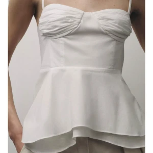 Vit volangtopp med smala axelband - Säljer en vit topp med smala axelband och volang nertill. Toppen har rynkade detaljer vid bysten och knytning i ryggen. Perfekt för en somrig look och har en feminin siluett. prislapp kvar.