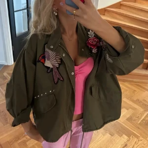 Olivgrön jacka med broderier från H&M - Säljer en populär olivgrön jacka från H&M med coola broderier. Jackan har oversized passform, lång ärm, tryckknappar och dekorativa nitar vid fickorna. Den är använd men inga defekter! 