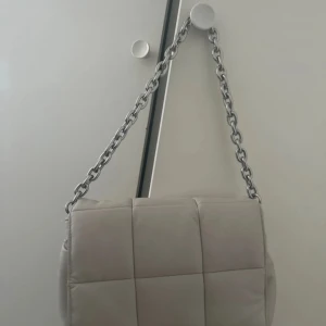 Beige quiltad axelväska med kedja - Säljer en beige quiltad axelväska med grov silverfärgad kedja som axelrem. Väskan har flera fack inuti och en mjuk, vadderad känsla. Perfekt för dig som vill ha en stilren och trendig accessoar.