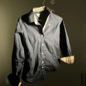 Mörkblå skjorta från Burberry - Säljer en mörkblå skjorta från Burberry med klassiska beige-rutiga detaljer på insidan av kragen och ärmsluten. Skjortan har knappar framtill och en broderad Burberry-logga på bröstet. Perfekt för dig som gillar stilrena plagg med lyxig känsla.