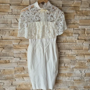 Vit spetsklänning med cape från Vesper - Supersnygg vit klänning från Vesper med elegant spetscape och rosett vid halsen. Klänningen är figurnära med hög hals och vackra spetsdetaljer upptill. Perfekt till studenten.