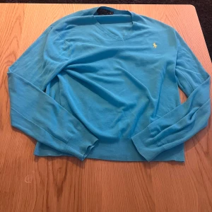 Turkos tröja från Polo Ralph Lauren - Säljer en turkos långärmad tröja från Polo Ralph Lauren i mjuk pima-bomull. Tröjan har v-ringad hals och den klassiska gula broderade loggan på bröstet. Perfekt för dig som gillar stilrena och färgglada plagg.