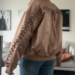 Beige bomberjacka med snörningsdetaljer - Säljer en beige bomberjacka med snygga snörningsdetaljer längs ärmarna och ryggen. Jackan har dragkedja framtill och är tillverkad i ett glansigt syntetmaterial. Perfekt för dig som vill ha en unik och trendig jacka.