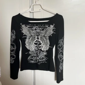 Svart långärmad topp med vingar och text - Säljer en svart långärmad topp med stort vitt tryck av änglavingar och text framtill och baktill. Ärmarna har också detaljerade vita mönster. Snygg och edgy stil, perfekt till jeans eller kjol.