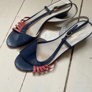 Blå och rosa sandaler med klack från S. Oliver - Säljer ett par stilrena sandaler från S. Oliver i blått skinn med rosa detaljer framtill. Skorna har öppen tå, låg klack och justerbart spänne vid ankeln. Perfekta för dig som vill ha en färgklick till outfiten!