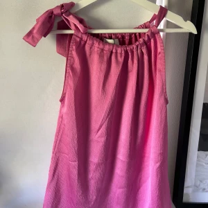 Rosa blus med knytband från Neo Noir - Säljer en söt rosa blus från Neo Noir med knytband på ena axeln. Blusen har en lös passform och är tillverkad i ett strukturerat tyg. Perfekt för varma dagar och somriga tillfällen.💐