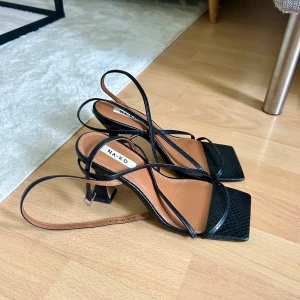 Svarta klackar från NA-KD - Säljer ett par svarta sandaler från NA-KD med smala remmar och fyrkantig tå. Skorna har en medelhög klack och är tillverkade i syntetmaterial med lätt ormmönstrad struktur. Perfekta för dig som gillar stilrena och trendiga skor.