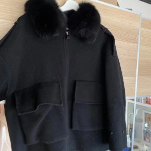 Svart ulljacka med äkta päls  - Säljer en svart jacka med dragkedja och stor, fluffig äkta päls runt kragen. Jackan har två stora fickor framtill och är långärmad. Perfekt för kyligare dagar. Puffarmar kommer med