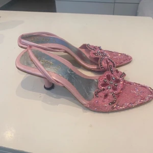 Rosa kitten heels med klack och pärldetaljer y2k - Säljer ett par rosa sandaler med öppen tå och klack. Skorna har dekorativa pärlor och paljetter framtill samt en elegant rosettdetalj. Perfekta för dig som vill sticka ut med glittriga detaljer och feminin stil. Skulle säga att det är 37or eller smala 38or. Något slitna
